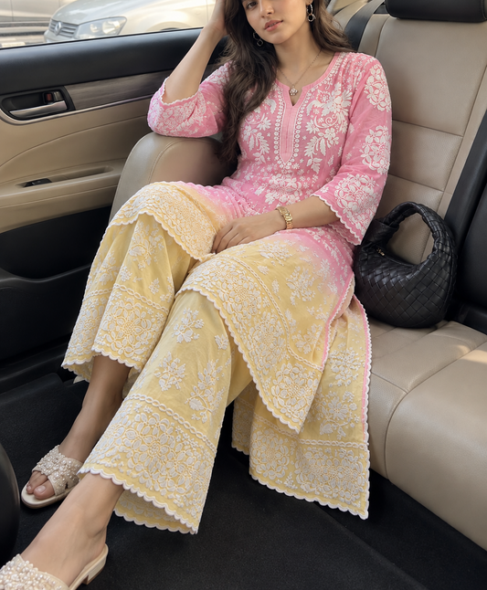 A woman seated in a beige car interior wearing a pink and pale yellow premium chikankari embroidered traditional outfit with white floral lace detailing. wearing chikankari kurti for women 