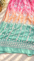 A close-up of a pastel gradient fabric, likely a traditional salwar suit material, featuring a transition from pink at the top to peach and mint green at the bottom. The fabric is decorated with intricate white floral embroidery and geometric borders.