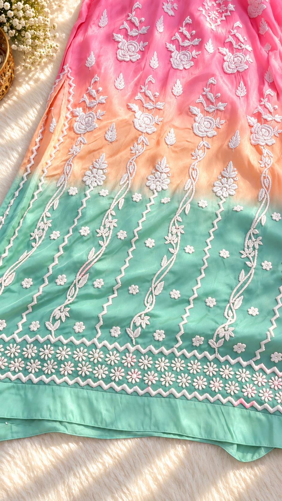A close-up of a pastel gradient fabric, likely a traditional salwar suit material, featuring a transition from pink at the top to peach and mint green at the bottom. The fabric is decorated with intricate white floral embroidery and geometric borders.