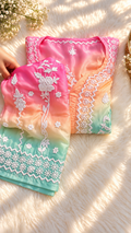 A folded pastel gradient garment with white floral embroidery, shown on a soft white faux-fur surface with warm sunlight and shadows. A hand is visible holding one corner of the fabric.