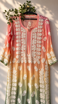 A three-quarter sleeve kurta with a peach, orange, and sage green ombre gradient, featuring intricate white floral embroidery down the front and on the sleeves. The kurta is displayed on a black hanger decorated with green leaves, set against a warm beige wall with soft leaf shadows.